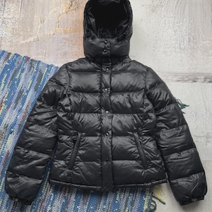 May include: A black puffer jacket featuring a quilted design, a hood, and a front closure with snap buttons. The jacket has long sleeves with elasticized cuffs and two side pockets. The material has a slight sheen, and the garment is fully padded.