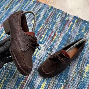 May include: A pair of dark brown leather shoes with a textured finish, featuring a moccasin-style stitched toe, a lace-up front with dark brown laces, and a low heel. Each shoe includes a metal shoe tree insert to maintain its shape.