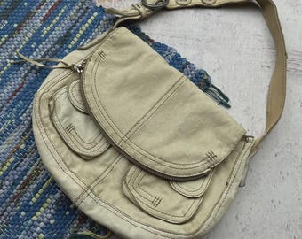May include: Cream-colored shoulder bag with a curved shape and multiple pockets. The bag features a zipper closure and a long, adjustable strap. Decorative stitching and metal hardware add detail to the design. The bag is made of a soft, textured material.