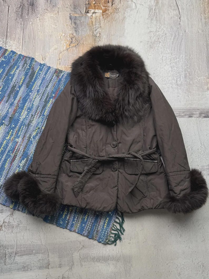 May include: A brown jacket with a fur collar and cuffs. The jacket has a belted waist and button closure. The fur is dark brown, and the jacket appears to be made of a quilted material. The jacket is displayed on a blue and white rug.