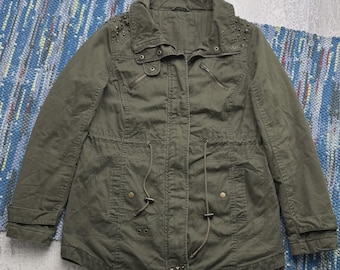 May include: An olive green utility jacket featuring a high collar, a front zipper closure with a snap-button placket, and long sleeves. The jacket includes two chest pockets, two waist pockets with drawstrings, and metallic stud embellishments.
