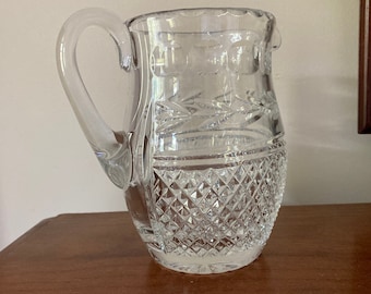 May include: A clear glass pitcher with a cut glass design. The pitcher has a handle and a wide mouth.