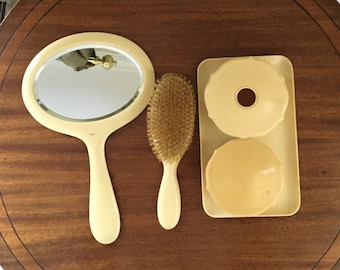 Può includere: Un set da toeletta vintage con uno specchio da mano bianco, una spazzola per capelli in legno e un contenitore a doppio scomparto con due coperchi rotondi color crema.