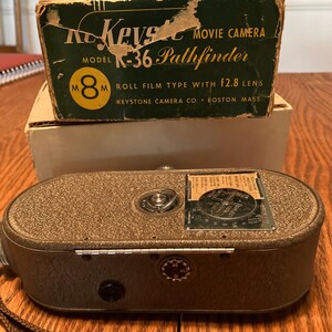 May include: A vintage Keystone K-36 Pathfinder 8mm movie camera in gray with a brown leather strap. The camera has a f2.8 lens and is in its original box.