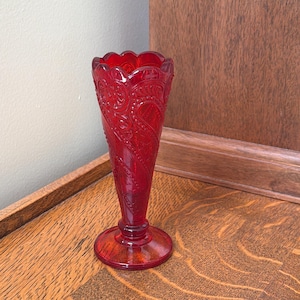 May include: A ruby red glass vase with a flared, scalloped rim. The vase features an embossed paisley design and a round base. The vase is approximately 8 inches tall.