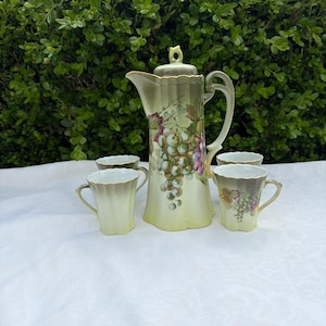 May include: A vintage tea set featuring a tall, light green coffee pot with a lid, and four matching cups. The coffee pot and cups are decorated with a grape cluster design in shades of purple, green, and brown. The set is on a white tablecloth.