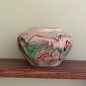 May include: A decorative, handcrafted vase with a unique marbled design. The vase features a blend of colors, including pink, green, orange, and black, creating a swirling pattern. The vase is sitting on a wooden shelf.