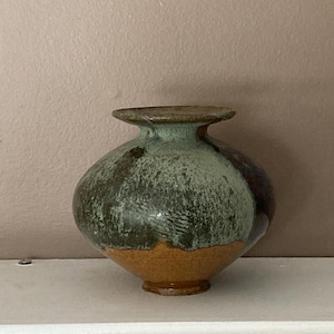 Può includere: Un vaso rotondo in ceramica con una base larga e piatta e un'apertura stretta. Il vaso è smaltato in tonalità di verde, marrone e arancio, con una finitura screziata e testurizzata.
