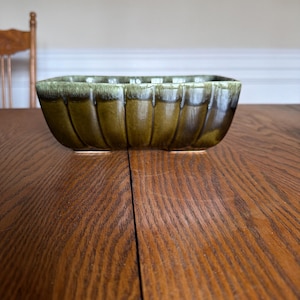 Puede incluir: Un cuenco rectangular de cerámica verde oliva con exterior acanalado. El cuenco tiene una superficie ligeramente texturizada y se apoya sobre dos pequeños pies. El borde tiene un esmalte más oscuro y moteado. El cuenco está sobre una mesa de madera.
