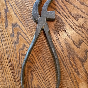 May include: A vintage rusty metal pliers with a hammer head on one side. The pliers are on a wooden surface.