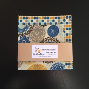 May include: A set of two beeswax food wraps in a medium size. The wraps feature a blue, brown, and yellow floral pattern. The packaging includes the text "Bienenwachstuch 2 tlg. Set "M" handgefertigt Die IdeenBiene www.ideenBiene.de"