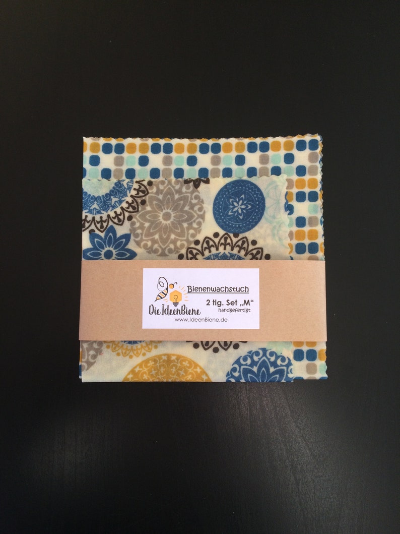 May include: A set of two beeswax food wraps in a medium size. The wraps feature a blue, brown, and yellow floral pattern. The packaging includes the text "Bienenwachstuch 2 tlg. Set "M" handgefertigt Die IdeenBiene www.ideenBiene.de"