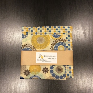 May include: Two sheets of beeswax food wrap with a blue, yellow, and brown geometric pattern. The wrap is labeled "Bienenwachstuch 2 flg. Set - 1" and "Die IdeenBiene handgefertigt www.ideenBiene.de".