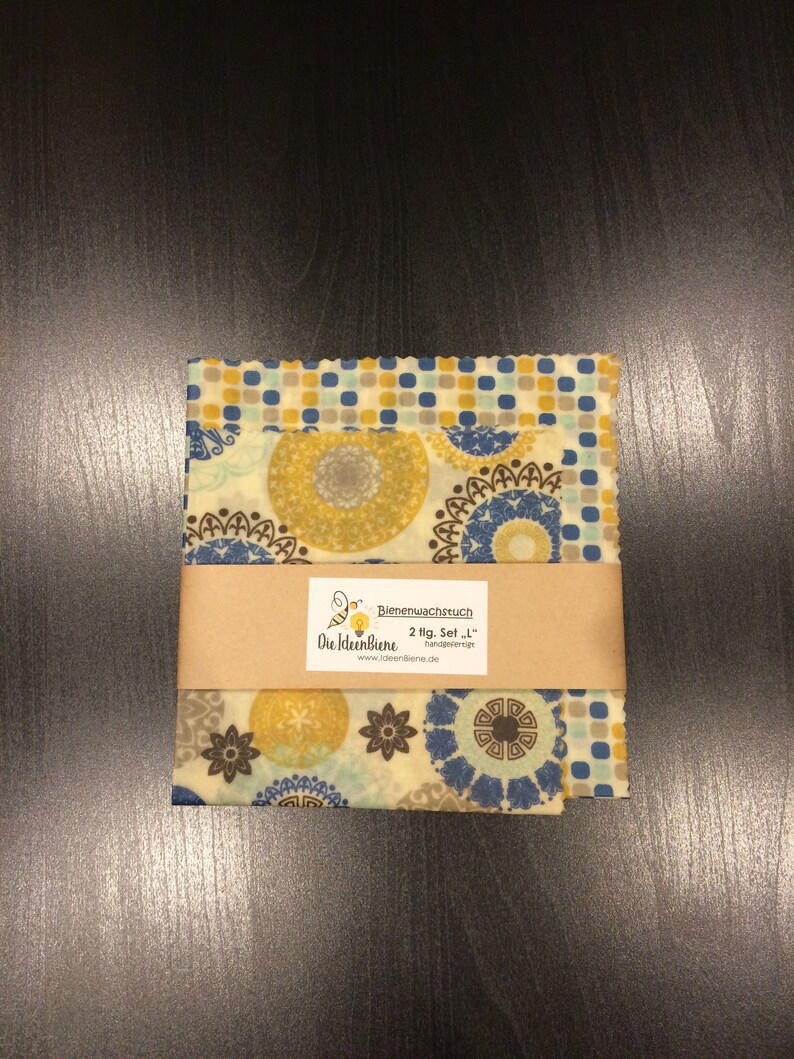 May include: Two sheets of beeswax food wrap with a blue, yellow, and brown geometric pattern. The wrap is labeled "Bienenwachstuch 2 flg. Set - 1" and "Die IdeenBiene handgefertigt www.ideenBiene.de".