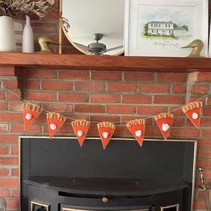 May include: A string of felt pumpkin pie slices hangs above a fireplace. The slices are orange with a white whipped cream topping. The fireplace is brick and has a black metal surround.