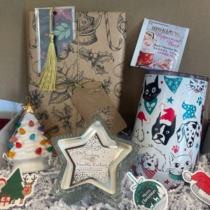 May include: A Christmas gift box filled with a variety of items, including a wrapped gift, a star-shaped candle, a ceramic Christmas tree ornament, a mug with a festive design, and a box of peppermint bark tea. The box is filled with white shredded paper.
