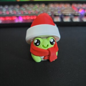 Könnte beinhalten: Eine grüne Froschfigur mit rotem Weihnachtsmannhut und Schal. Der Frosch hat große, ausdrucksstarke Augen und einen lächelnden Mund. Der Hut hat eine weiße Krempe und der Schal ist um den Hals des Frosches gewickelt. Die Figur ist wahrscheinlich eine Weihnachtsdekoration.
