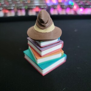 May include: A miniature brown witch hat with a tan band sits atop a stack of colorful books. The books are teal, orange, purple, and white, with pink and white edges. The objects are likely 3D printed.