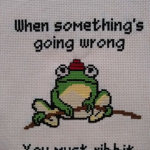 May include: Cross-stitch embroidery of a green frog wearing a red hat, sitting on a branch. The text reads "When something's going wrong, you must ribbit."