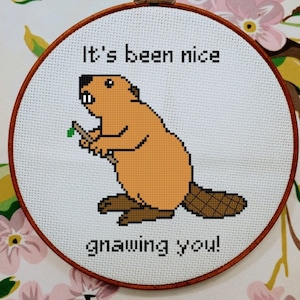 May include: A cross-stitch embroidery hoop with a brown beaver holding a stick and the text "It's been nice gnawing you!"