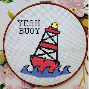 Pode incluir: Um aro de bordado de ponto cruz branco com uma bóia vermelha e branca com uma parte superior amarela. A bóia está em ondas azuis. O texto "YEAH BUOY" é bordado em preto acima da bóia.