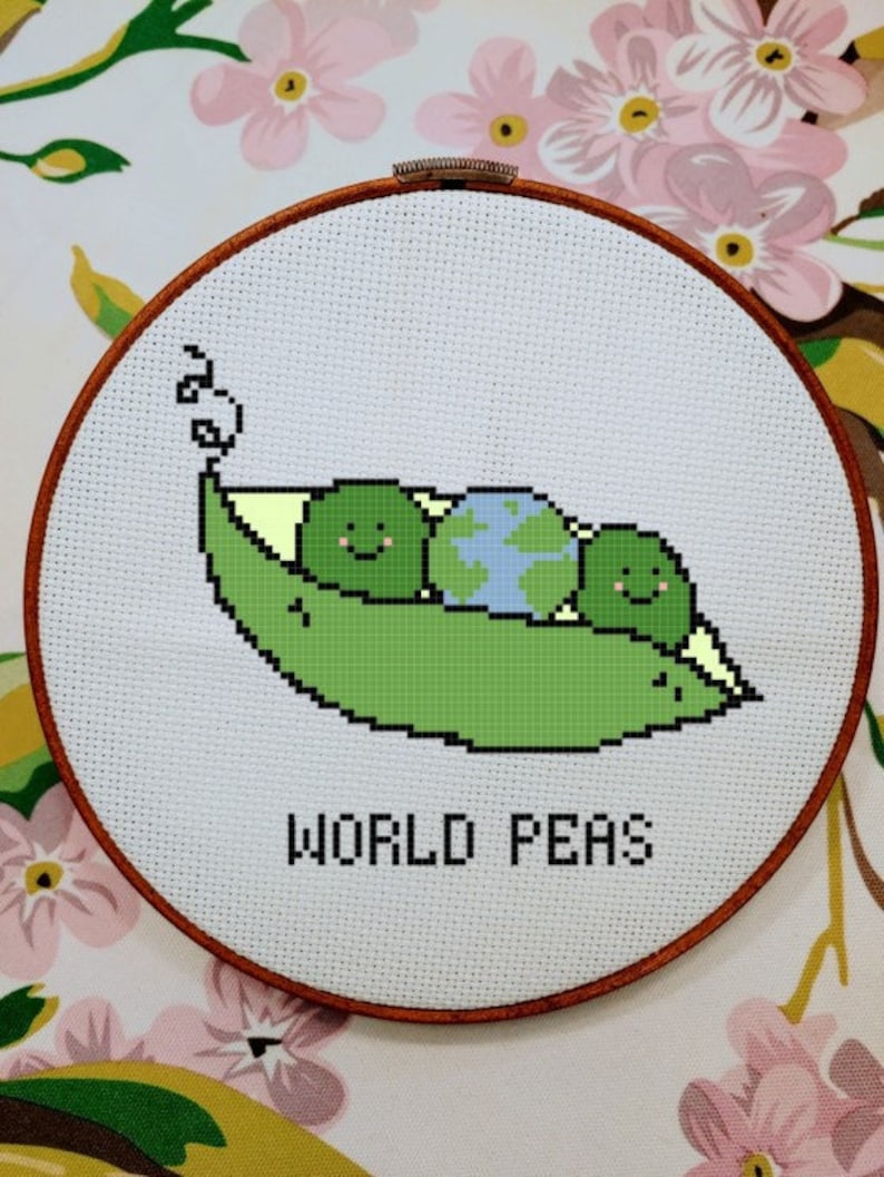 May include: A white embroidery hoop with a green pea pod containing three green peas and a blue and green globe. The text "WORLD PEAS" is embroidered below the pea pod.