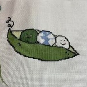 May include: Cross-stitch embroidery of three green peas in a pea pod. The peas have smiling faces and one has a blue and white pattern.