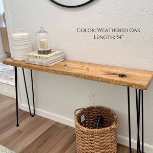 Puede incluir: Una mesa consola de roble envejecido con patas de horquilla negras. La mesa tiene 137 cm de largo y un dise&ntilde;o de borde vivo. Est&aacute; decorada con un jarr&oacute;n blanco, una campana de cristal con una vela y una pila de libros.