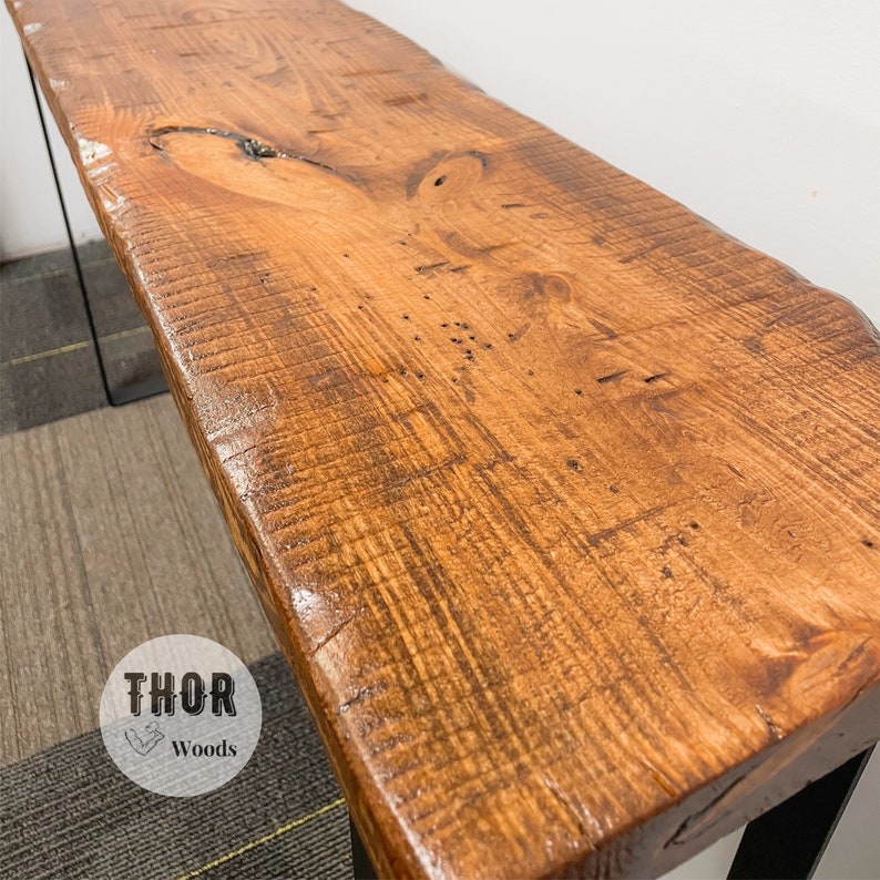 May include: A rustic wooden console table with a live edge top and black metal legs. The table has a natural wood finish and is made from reclaimed wood. The table is approximately 48 inches long and 16 inches wide.