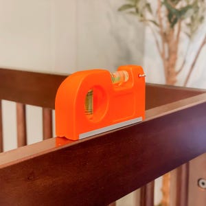 May include: Bright orange level tool with a clear vial containing a green liquid. The level is resting on a dark brown wooden surface. The tool has a silver base and a hole in the center.