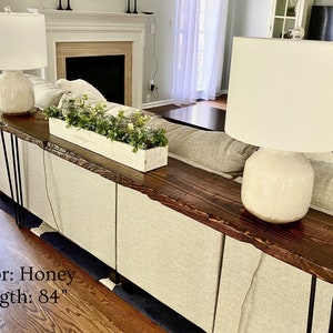 Puede incluir: Una mesa consola de madera de color miel con patas de horquilla de metal negro. La mesa tiene 84 pulgadas (213 cm) de largo y presenta un dise&ntilde;o de borde vivo. Est&aacute; decorada con dos l&aacute;mparas de cer&aacute;mica blanca y una maceta con vegetaci&oacute;n.