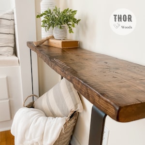 May include: A rustic wooden console table with a live edge top and black metal legs. The table is decorated with a potted plant, a book, and a decorative tassel. The table is labeled "THOR Woods".