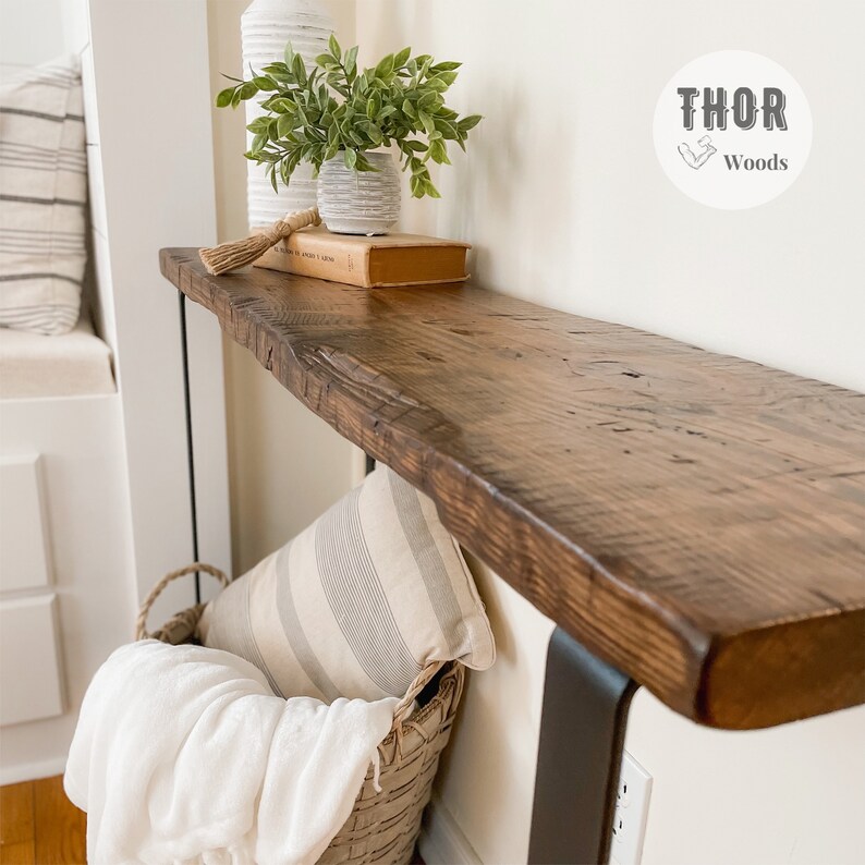 May include: A rustic wooden console table with a live edge top and black metal legs. The table is decorated with a potted plant, a book, and a decorative tassel. The table is labeled "THOR Woods".