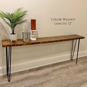 Puede incluir: Una mesa consola de nogal de 183 cm de largo con una tapa de borde vivo y patas de horquilla negras. La mesa est&aacute; decorada con una planta en maceta, una vela y una letra decorativa U.