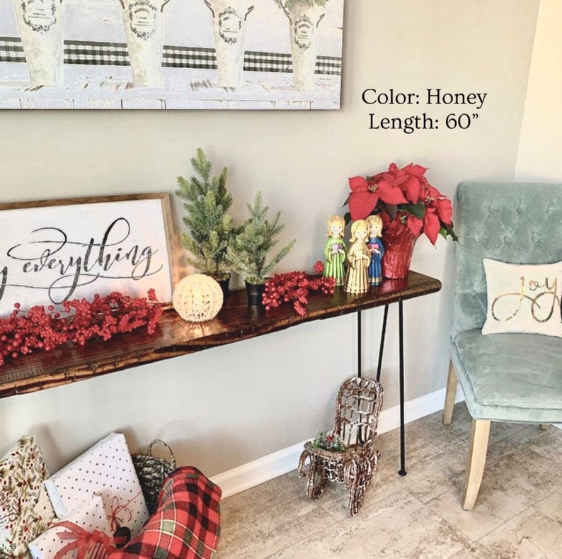 Puede incluir: Una mesa consola de madera color miel con patas de horquilla de metal negro. La mesa est&aacute; decorada con un letrero que dice "Joy everything", dos peque&ntilde;os &aacute;rboles de Navidad en maceta, ramitas de bayas rojas y una planta de poinsettia en una maceta roja. Dos peque&ntilde;as figuras de &aacute;ngeles est&aacute;n de pie sobre la mesa. La mesa tiene 152,4 cm de largo.