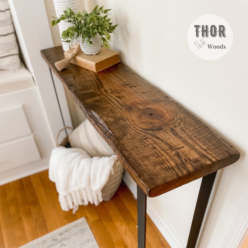 May include: A rustic wooden console table with a live edge top and black metal legs. The table is decorated with a potted plant, a book, and a woven basket with a white blanket.