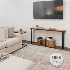 May include: A wooden console table with black metal legs, featuring a rustic wood top and two woven baskets filled with blankets. The table is placed in front of a white wall with a flat screen TV mounted above it. The text "THOR Woods" is visible in the lower right corner of the image.
