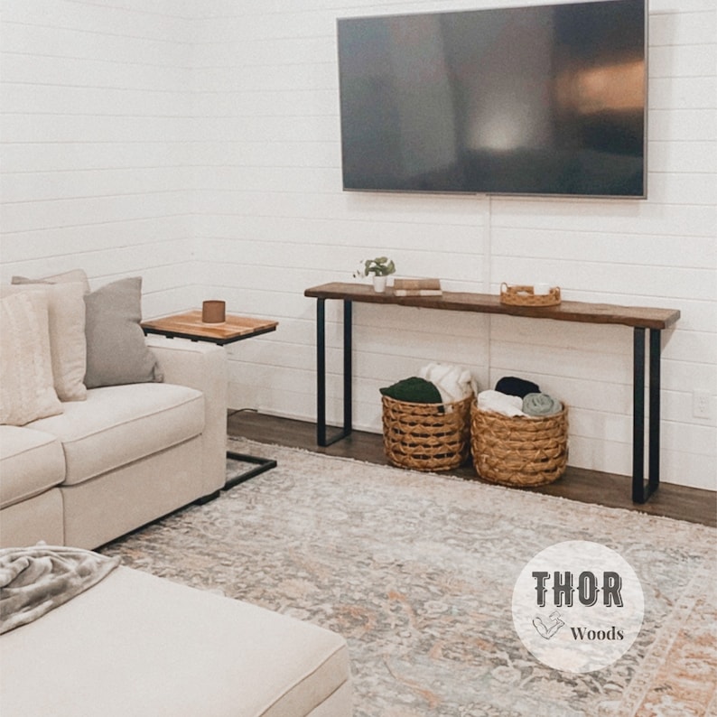 May include: A wooden console table with black metal legs, featuring a rustic wood top and two woven baskets filled with blankets. The table is placed in front of a white wall with a flat screen TV mounted above it. The text "THOR Woods" is visible in the lower right corner of the image.