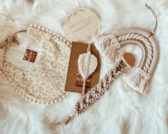 May include: A set of baby items in a neutral color palette. The set includes a beige bib with a floral pattern, a wooden sign that says "Hello World", a macrame rainbow with a wooden teether, and a small book with a feather bookmark.