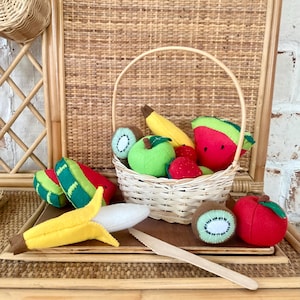 Könnte beinhalten: Ein Filz-Obst-Spielset mit einer Auswahl an Früchten, darunter eine Banane, ein Apfel, eine Kiwi, eine Wassermelone und eine Erdbeere. Die Früchte befinden sich in einem Weidenkorb mit einem Holzgriff. Außerdem gibt es ein Holzmesser und ein Holzbrett.