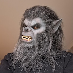 May include: A werewolf mask with grey fur and a snarling expression. The mask features a detailed face with sharp teeth, pointed ears, and a long beard. The mask is worn over a black hooded sweatshirt.