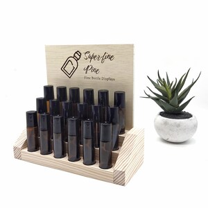May include: A wooden display stand with multiple amber glass roller bottles with black caps. The stand has a light wood finish and a wooden sign that reads "Super-fine Pine Fine Bottle Displays". A small potted succulent is to the right.