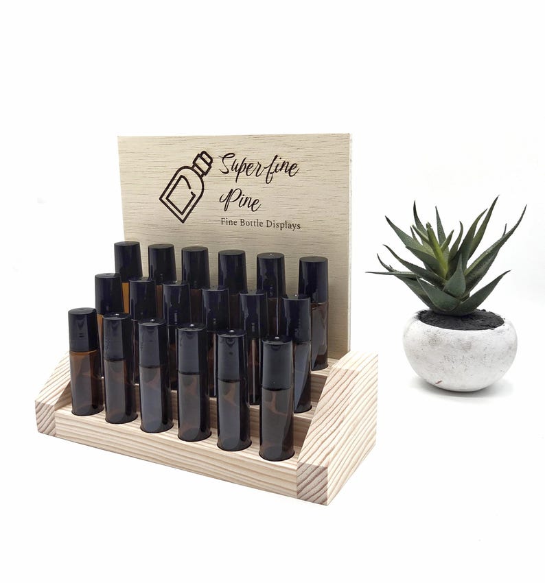 May include: A wooden display stand with multiple amber glass roller bottles with black caps. The stand has a light wood finish and a wooden sign that reads "Super-fine Pine Fine Bottle Displays". A small potted succulent is to the right.