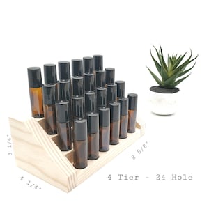 May include: A wooden essential oil display stand with 24 holes, arranged in four tiers. The stand holds numerous amber glass roller bottles with black caps. A small succulent plant in a white pot is in the background. The stand measures 8 5/8" long, 4 1/4" wide, and 3 1/4" high.