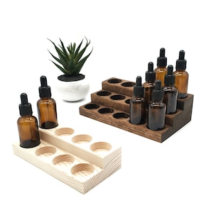 May include: Two tiered wooden display stands for essential oil bottles. One stand is light natural wood, the other is dark stained wood. Both feature circular cutouts for amber glass dropper bottles with black caps. A small green succulent is included.