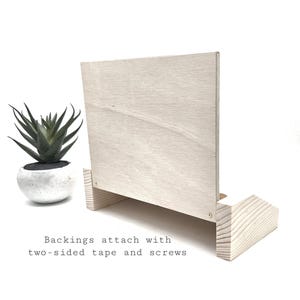 May include: A light-colored wooden display stand with a rectangular back and a base. The text "Backings attach with two-sided tape and screws" is visible. A small potted succulent is to the left.