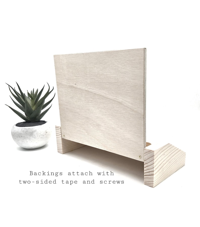 May include: A light-colored wooden display stand with a rectangular back and a base. The text "Backings attach with two-sided tape and screws" is visible. A small potted succulent is to the left.