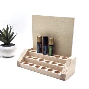 May include: A tiered, unfinished wooden essential oil display stand with three rows of holes. Three essential oil bottles are displayed on the top tier. A small potted succulent is to the left of the stand.