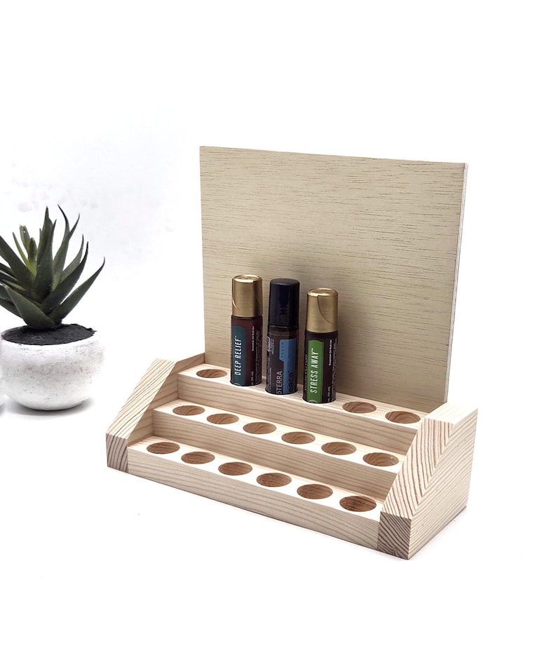 May include: A tiered, unfinished wooden essential oil display stand with three rows of holes. Three essential oil bottles are displayed on the top tier. A small potted succulent is to the left of the stand.