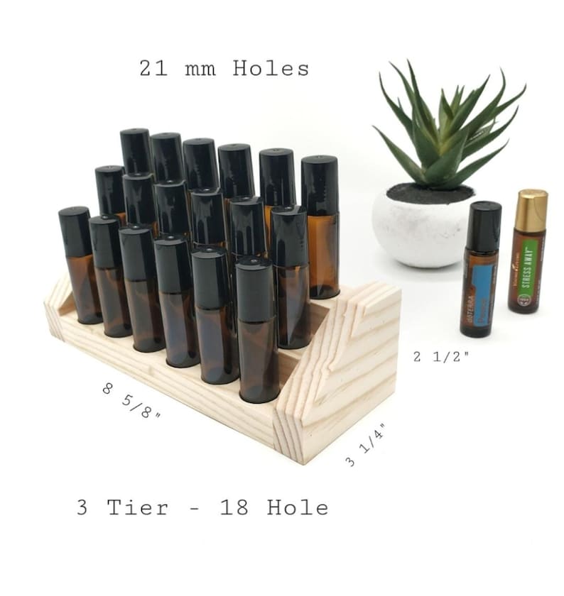 May include: A three-tiered, 18-hole wooden essential oil display stand. The stand holds several amber glass roller bottles with black caps. The stand measures 8 5/8" long, 3 1/4" wide, and 2 1/2" tall. The holes are 21 mm.